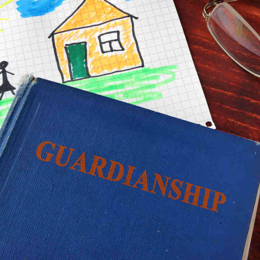 A blue book titled "GUARDIANSHIP" rests on a wooden table, next to a child's drawing of a family and house, and a pair of eyeglasses, symbolizing the legal aspects of guardianship.